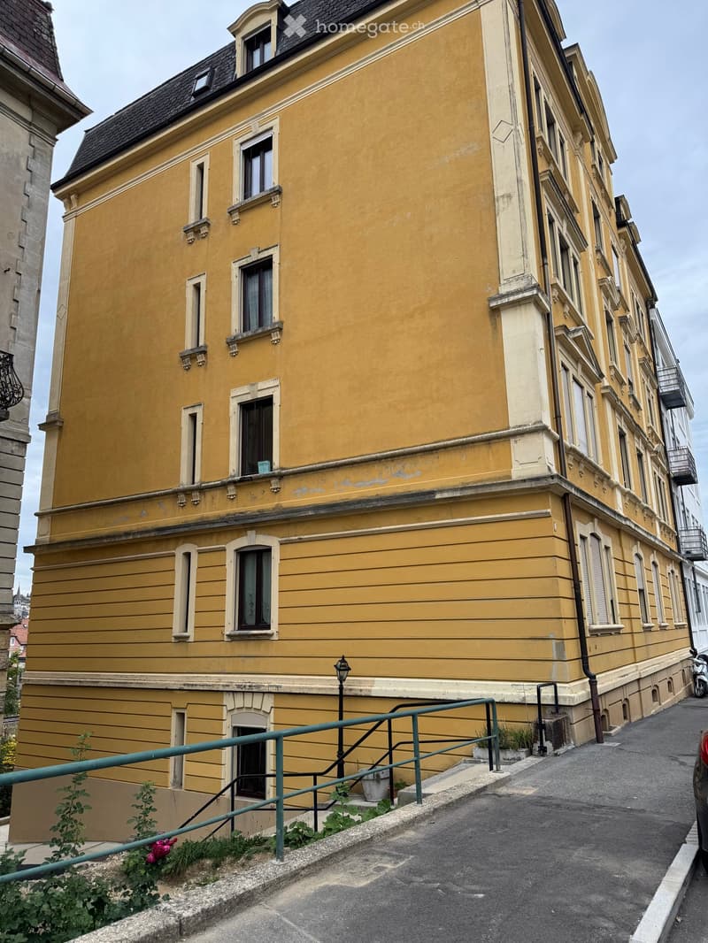 A multi-story yellow building with classic architectural details, including ornate window frames and decorative moldings. The building has multiple windows and a small staircase leading to the entrance.
