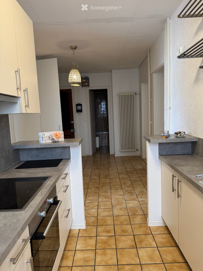 Modern kitchen with white cabinets, grey countertops, induction stove, oven, and a small seating area with a cup and flower on the counter.