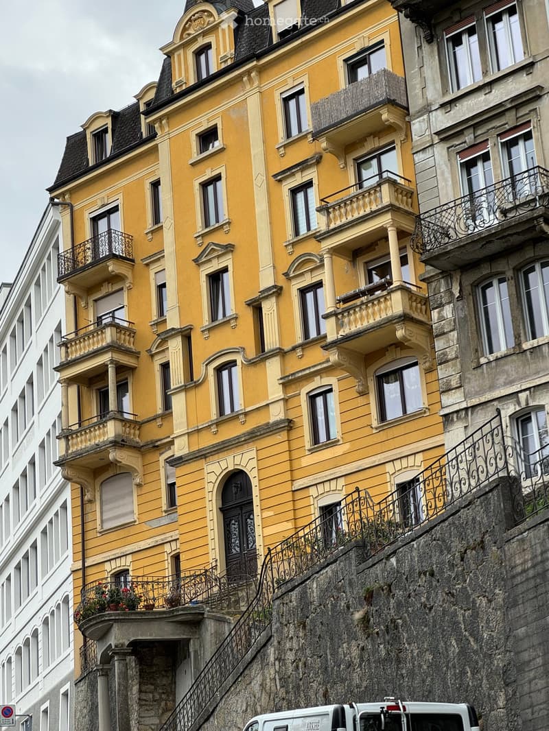 A multi-story yellow building with ornate balconies and decorative railings, featuring multiple windows and a stone staircase leading up to the entrance.