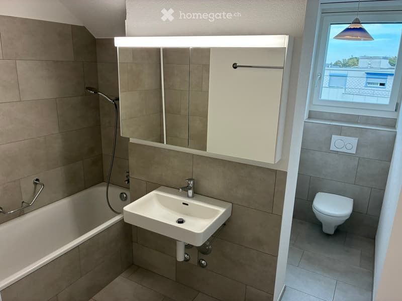 Modern bathroom with a bathtub, sink, toilet, mirror, and built-in cabinet. Features tiled walls and a window with a view.