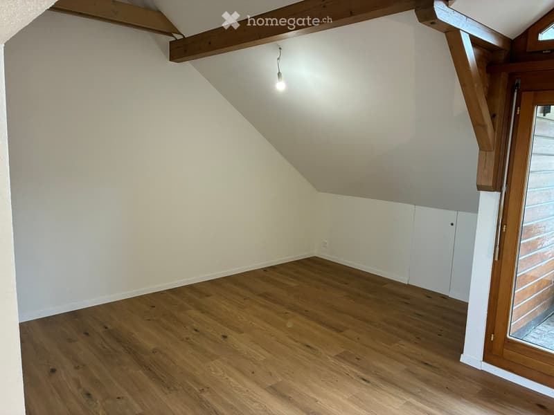 Empty attic room with wooden flooring, white walls, exposed wooden beams, and a single hanging light fixture.