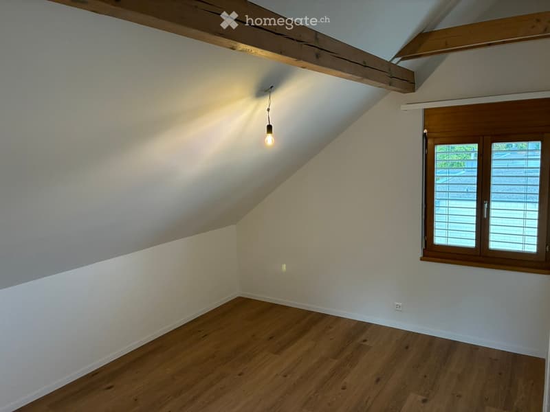 Empty attic room with wooden floor, exposed wooden beams, and a single window with blinds. The room has white walls and a hanging light fixture.
