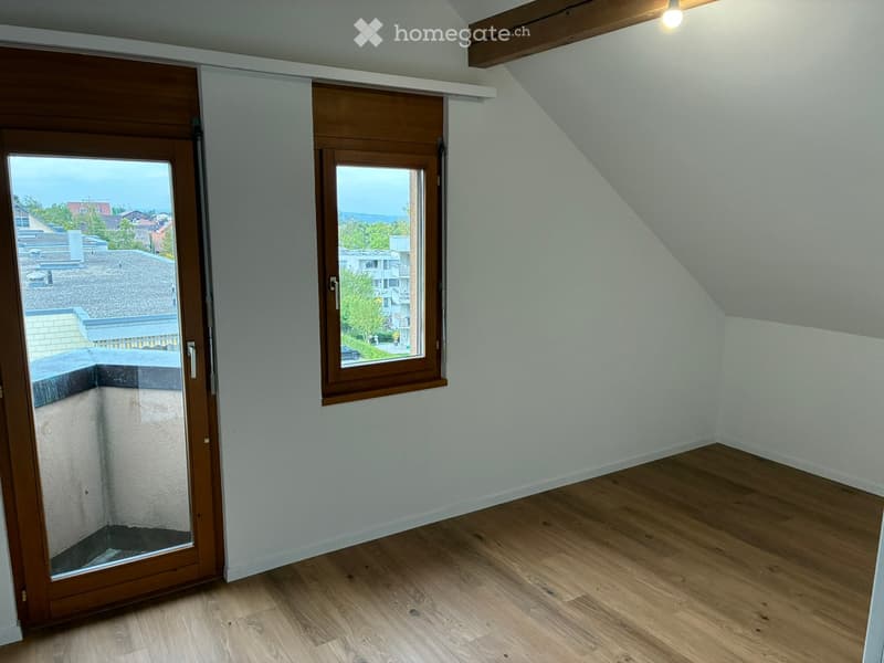 Empty attic room with wooden flooring, white walls, and large windows offering a view of the neighborhood.