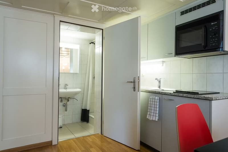 A small, modern kitchen with a microwave, sink, and faucet, connected to a compact bathroom with a shower and sink. A red chair is visible near the kitchen counter.