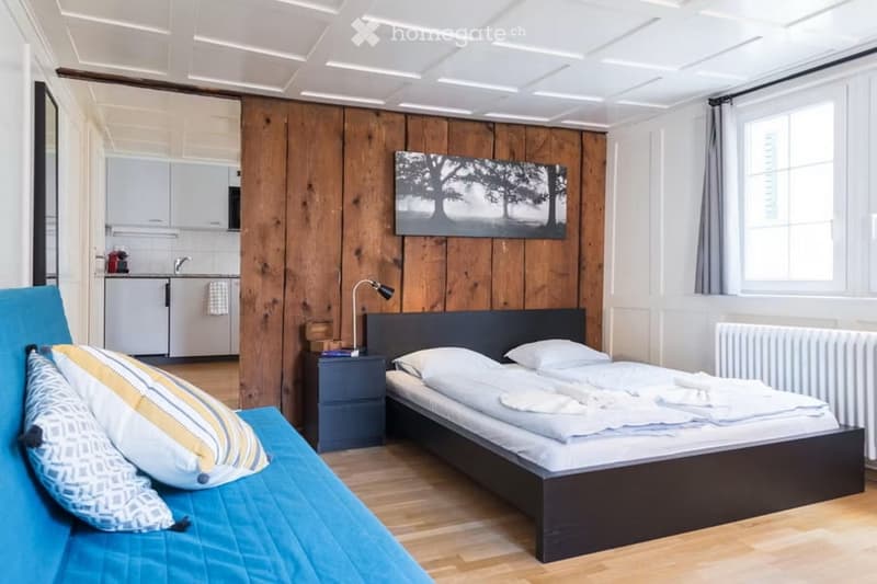 A modern, compact bedroom with a double bed, nightstand, sofa, and a small kitchen area with cabinets and a sink.
