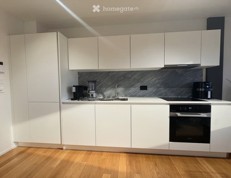 Modern kitchen with white cabinets, marble backsplash, and wooden flooring. Appliances include a coffee maker, blender, and oven.