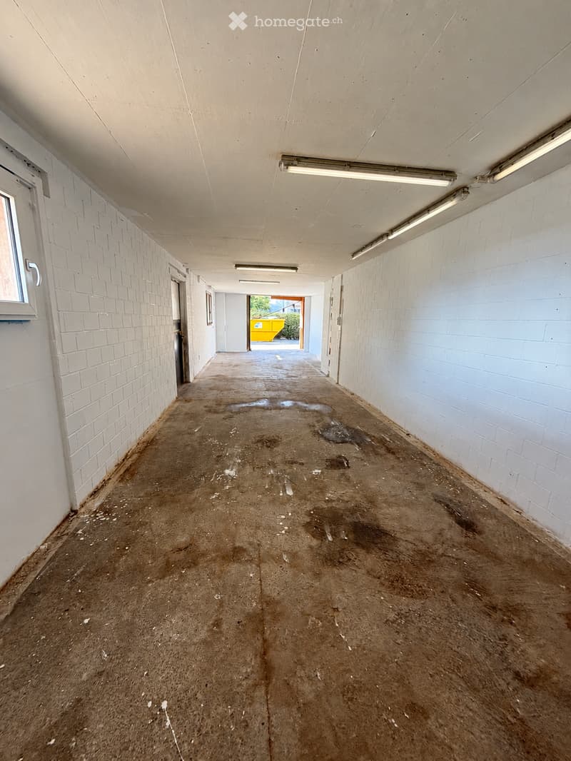 Empty corridor with white brick walls, concrete floor, and fluorescent lighting. The floor shows signs of wear and dirt, with a small puddle visible.