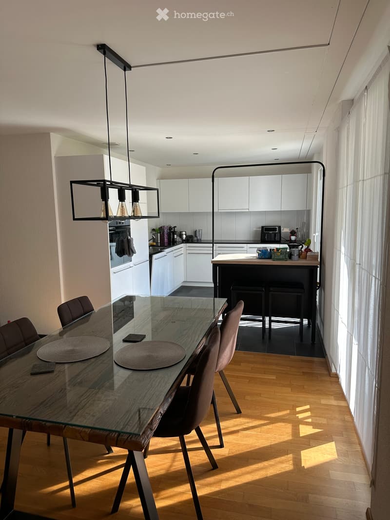 Modern open-plan dining and kitchen area with a wooden dining table, glass top, and chairs. Kitchen features white cabinets, stainless steel appliances, and a central island with bar stools.