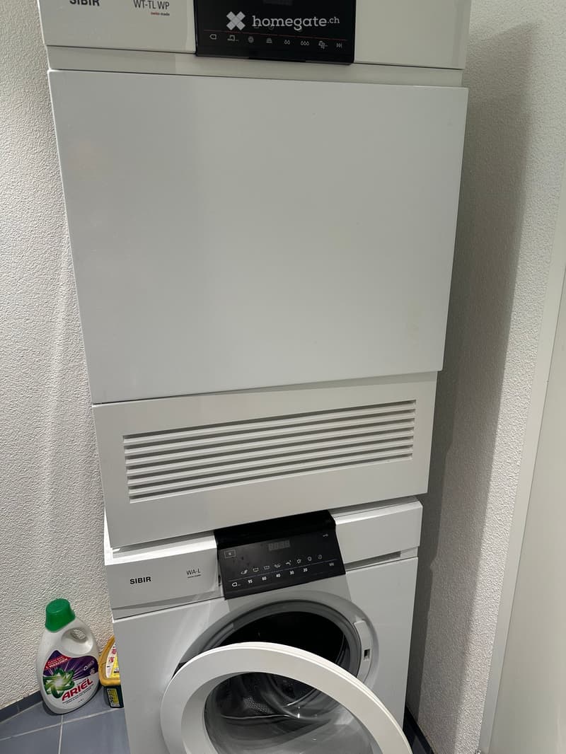 A compact bathroom setup featuring a stacked washing machine and dryer unit with control panels, accompanied by a bottle of detergent placed on the floor.