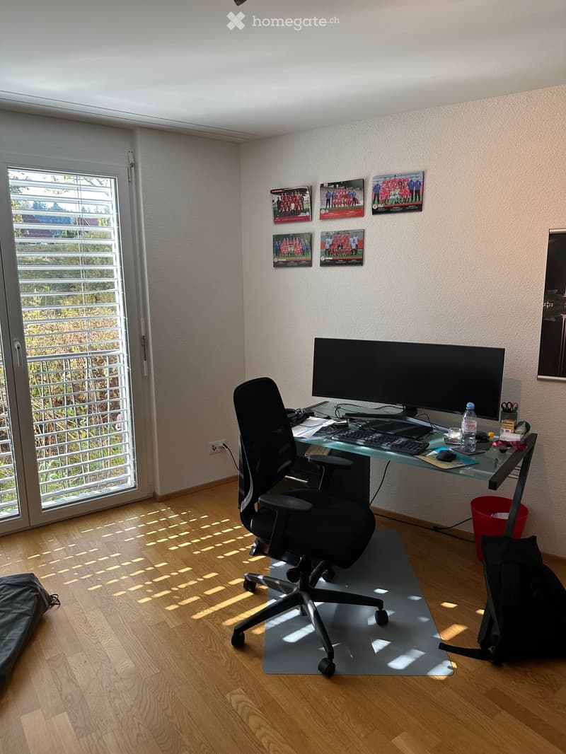 A modern, well-lit room with a wooden floor, a large window with blinds, a desk with a screen, keyboard, mouse, and a chair, along with posters on the wall and a red waste bin.