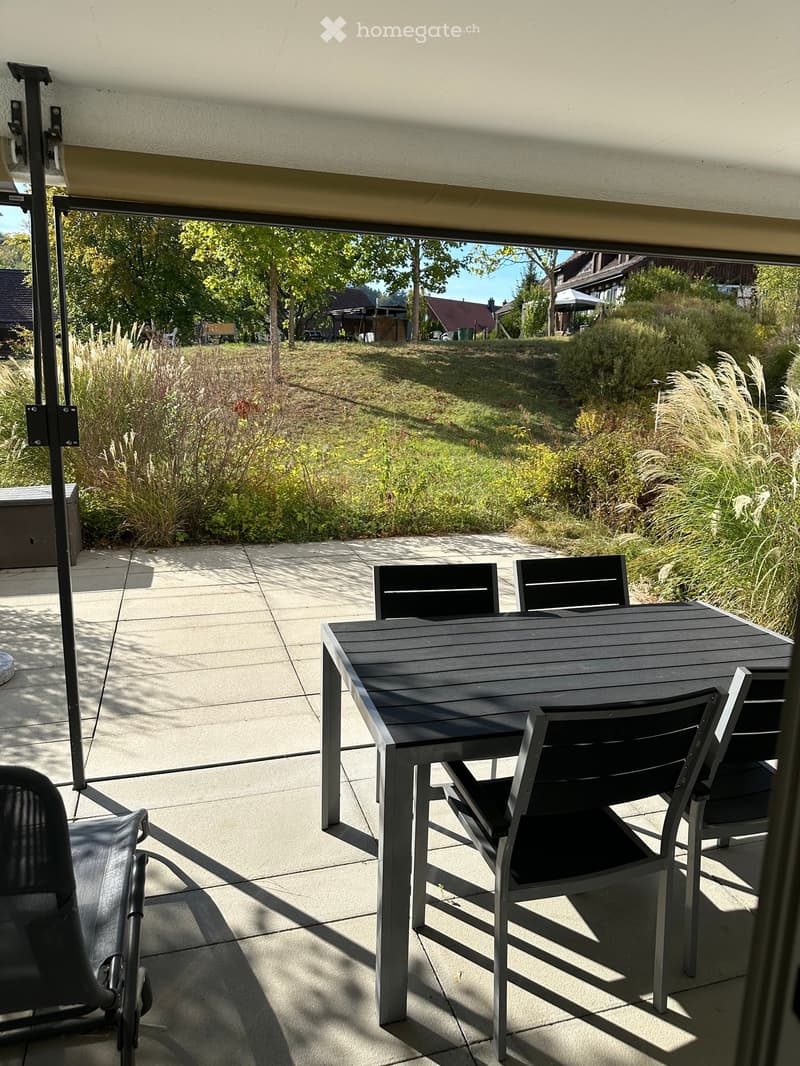 Outdoor patio with a black table and chairs, surrounded by greenery and a view of a garden and trees.