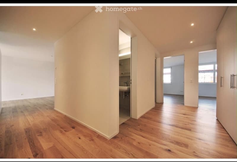 Empty hallway with wooden flooring and white walls, leading to a small bathroom and another room with large windows.