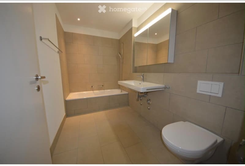 Modern bathroom with beige tiles, a bathtub, a sink, a toilet, and a large mirror with built-in lighting.
