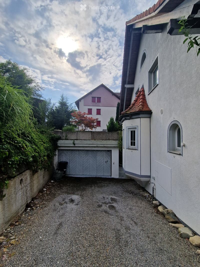 Two-story house with white exterior, red-tiled roof, garage, and surrounding greenery.
