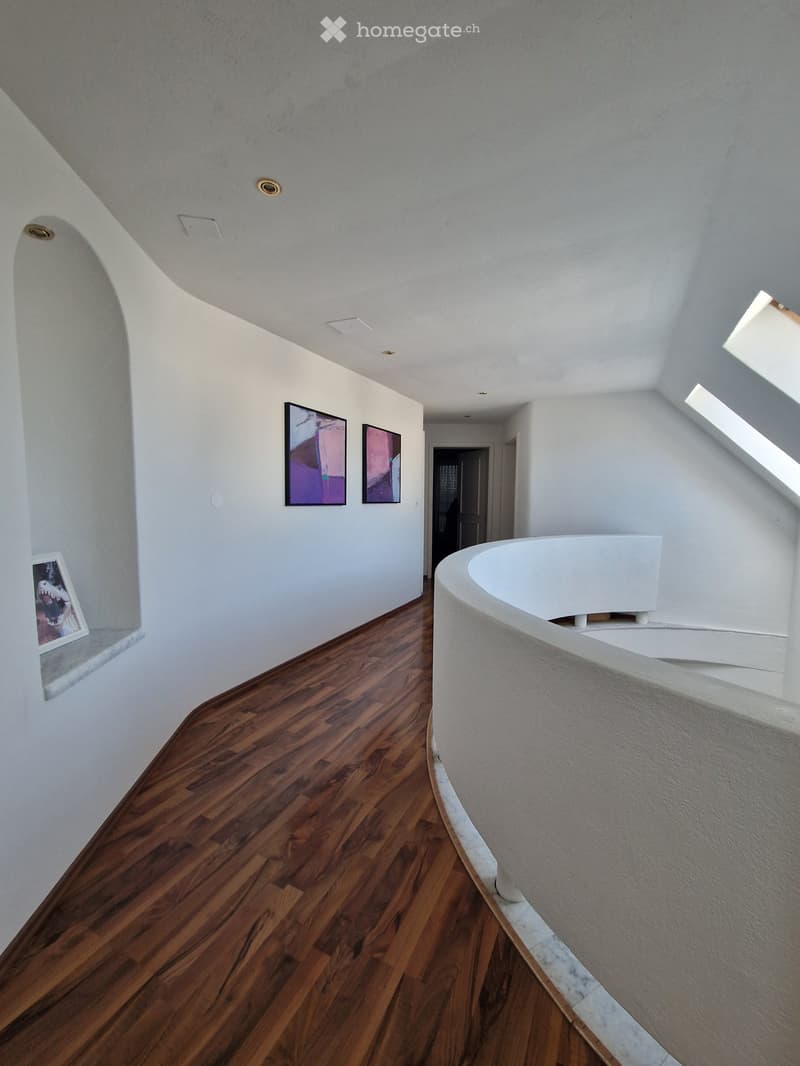 Spacious hallway with wooden flooring, white walls, and modern lighting fixtures. Features a curved white staircase railing and several framed artworks on the walls.