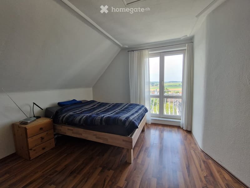 Bedroom with wooden floor, slanted ceiling, large window with white curtains, bed with blue bedding, nightstand with lamp and radio.