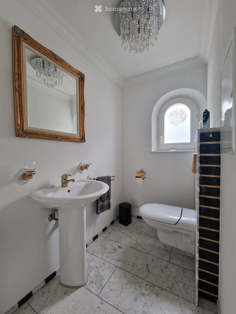 Elegant bathroom with a white pedestal sink, a toilet, a large ornate mirror, a chandelier, and a window with an arched design.