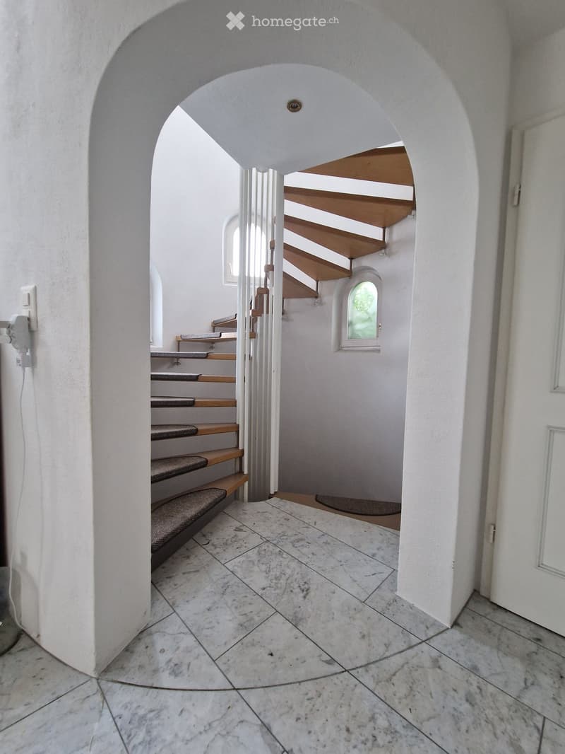 A bright, arched corridor with marble flooring and a staircase leading upwards, featuring wooden steps and a small window on the wall.