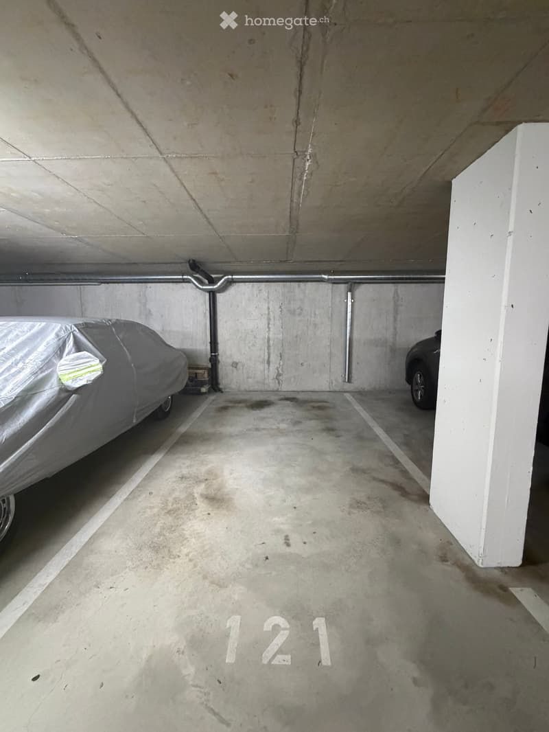 Underground parking space with concrete walls and ceiling, marked with number "121". Two cars are parked, one covered with a tarp.