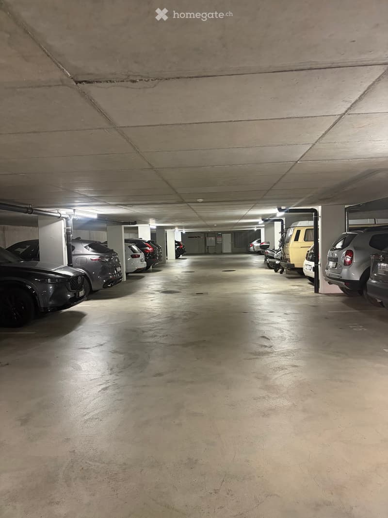 Underground parking area with several parked cars and a motorcycle, concrete floors and walls, fluorescent lighting.