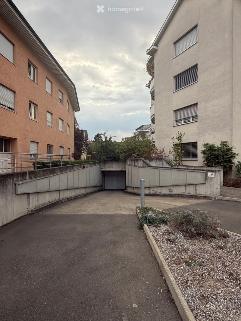 A paved driveway leading to a garage entrance, flanked by modern apartment buildings with balconies and windows.