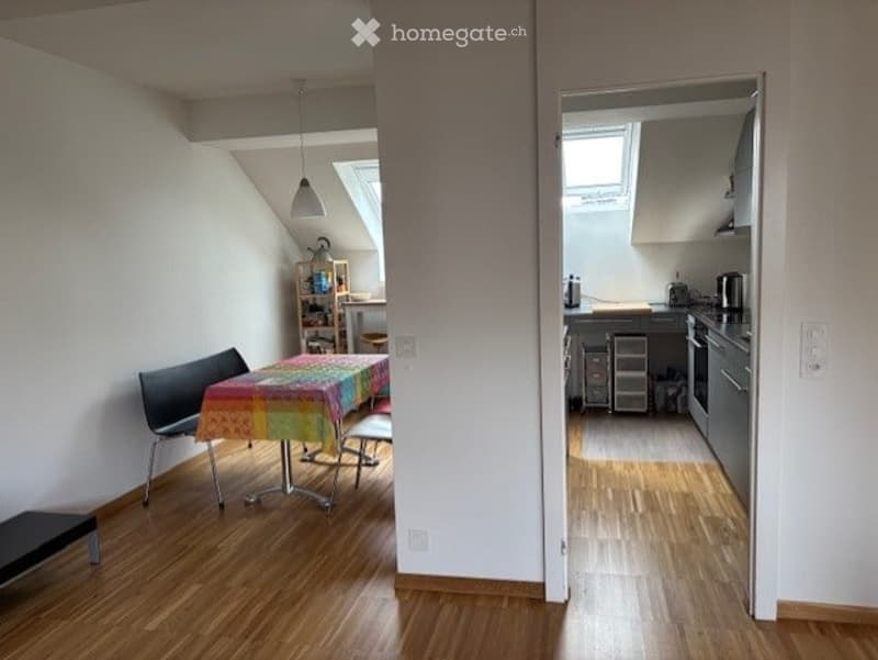 The image shows a modern, open-plan living and dining area connected to a small kitchen. The living area features a wooden floor, a colorful tablecloth-covered dining table with chairs and a bench, and a bookshelf with various items. The kitchen area incl