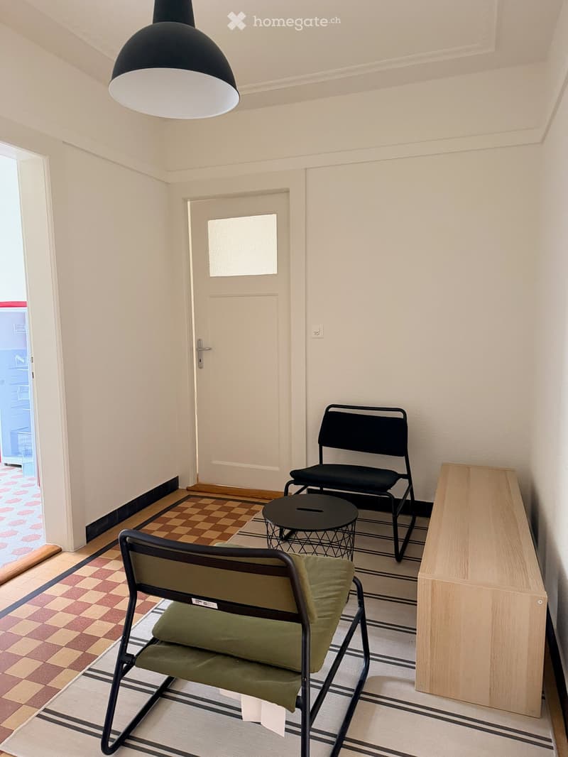 A small, furnished room with a wooden floor, a black hanging lamp, a green chair, a black chair, a small round table, and a wooden cabinet.