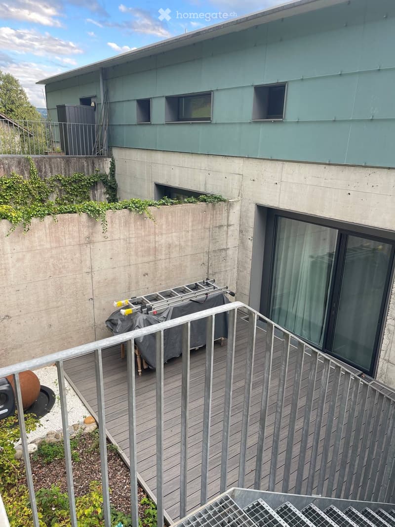 Balcony with wooden decking, metal railing, covered furniture, and a view of a modern building with green walls and concrete base.