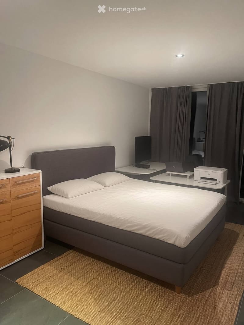 A modern bedroom with a double bed, nightstand, lamp, laptop, printer, and a large window with dark curtains.