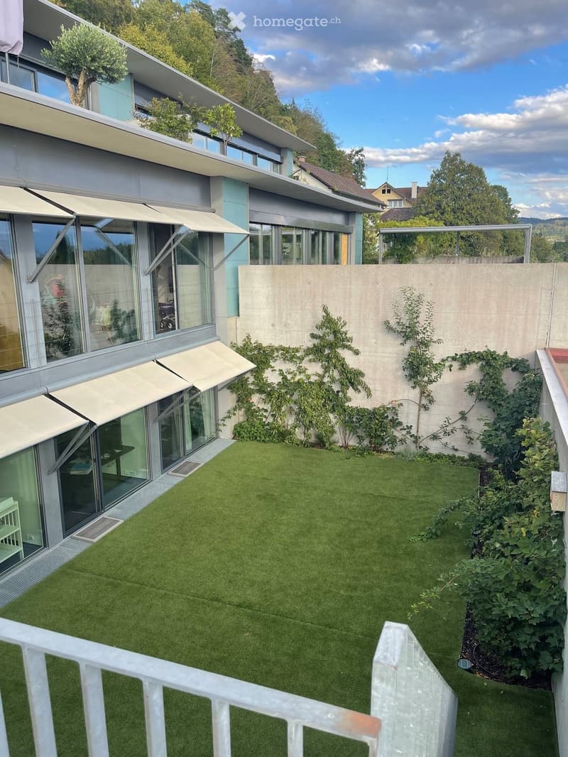Modern house with a well-maintained artificial grass lawn, surrounded by climbing plants and shrubs. The house features large glass windows and sliding doors, with a concrete wall and small trees in the background.