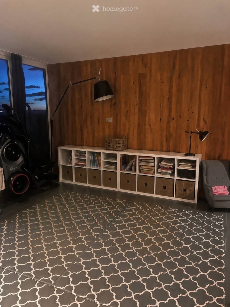Modern living room with wooden wall paneling, grey carpet with geometric pattern, white bookshelf filled with books and storage boxes, grey armchair, floor lamp, table lamp, and exercise bike near the window.