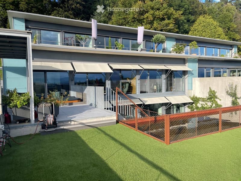 Modern two-story house with large windows, balconies, and a well-maintained lawn. Features include a staircase leading to the upper level and outdoor furniture on the deck.