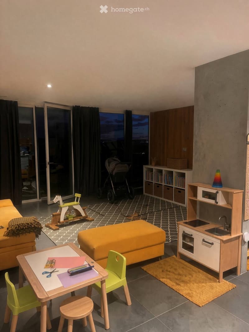 A modern, well-lit living space with a sofa, a small bed, a play kitchen, a bookshelf, and various toys including a toy horse and a stroller. The room features large windows with a view and a cozy rug.