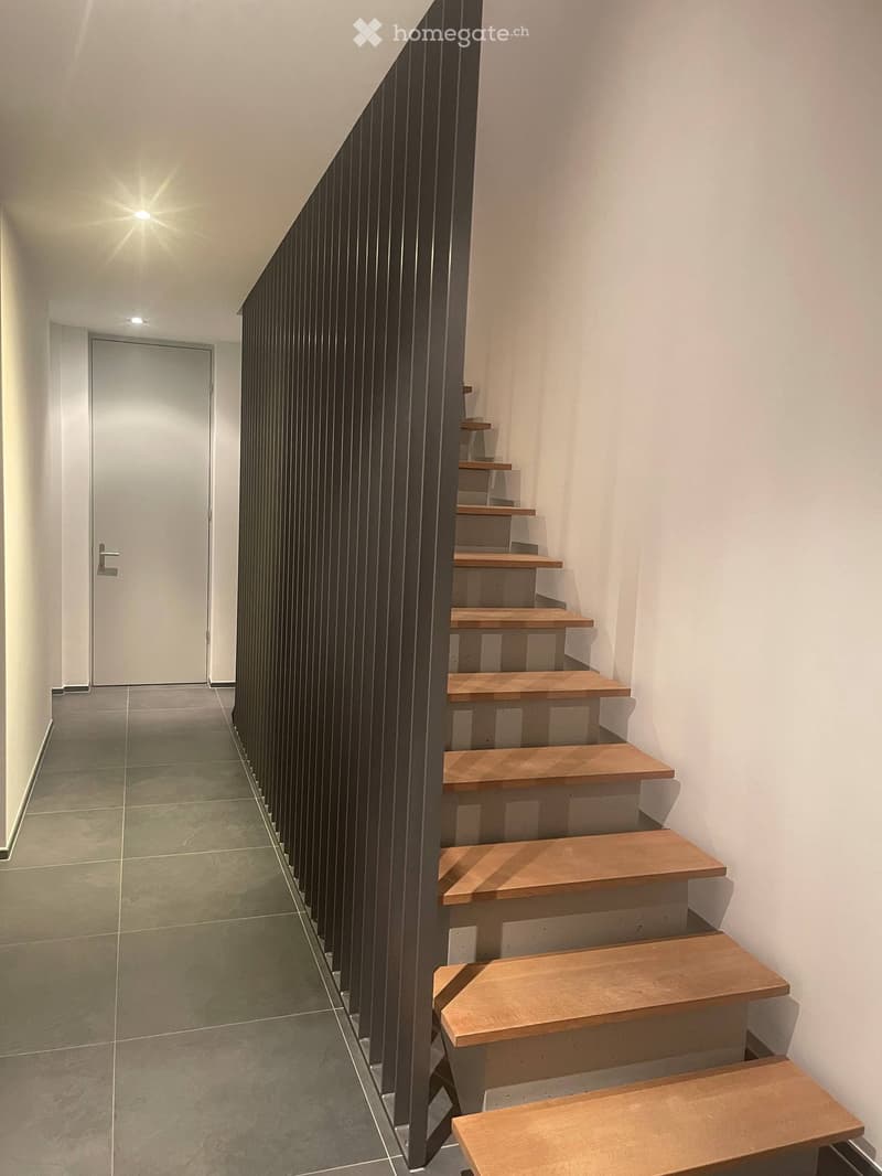 Modern staircase with wooden steps and a sleek black railing, leading up to a door at the end of a tiled hallway.