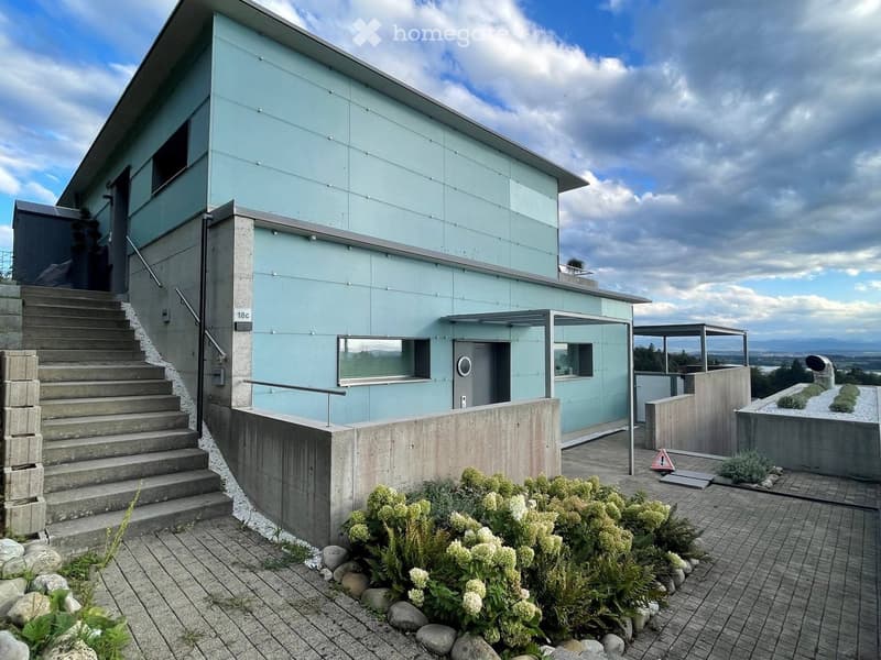 Modern two-story building with light blue exterior, concrete steps leading to entrance, small garden area with shrubs and rocks, and a view of the landscape.