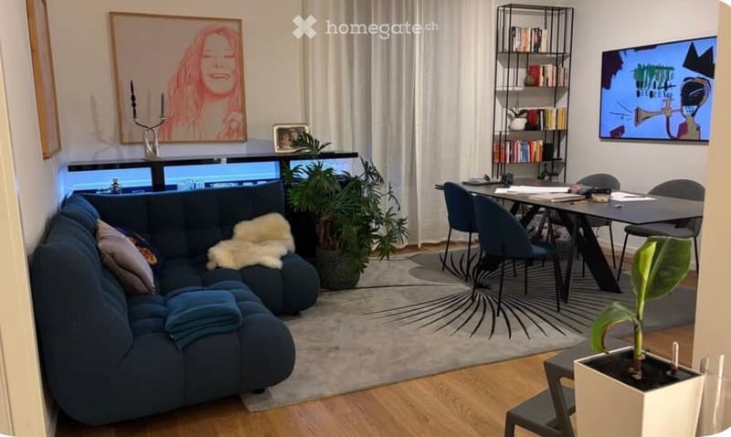 A modern living room with a blue sectional sofa, a dining table with chairs, a large plant, and a TV screen. The room features wooden flooring, a large rug, and decorative artwork on the walls.