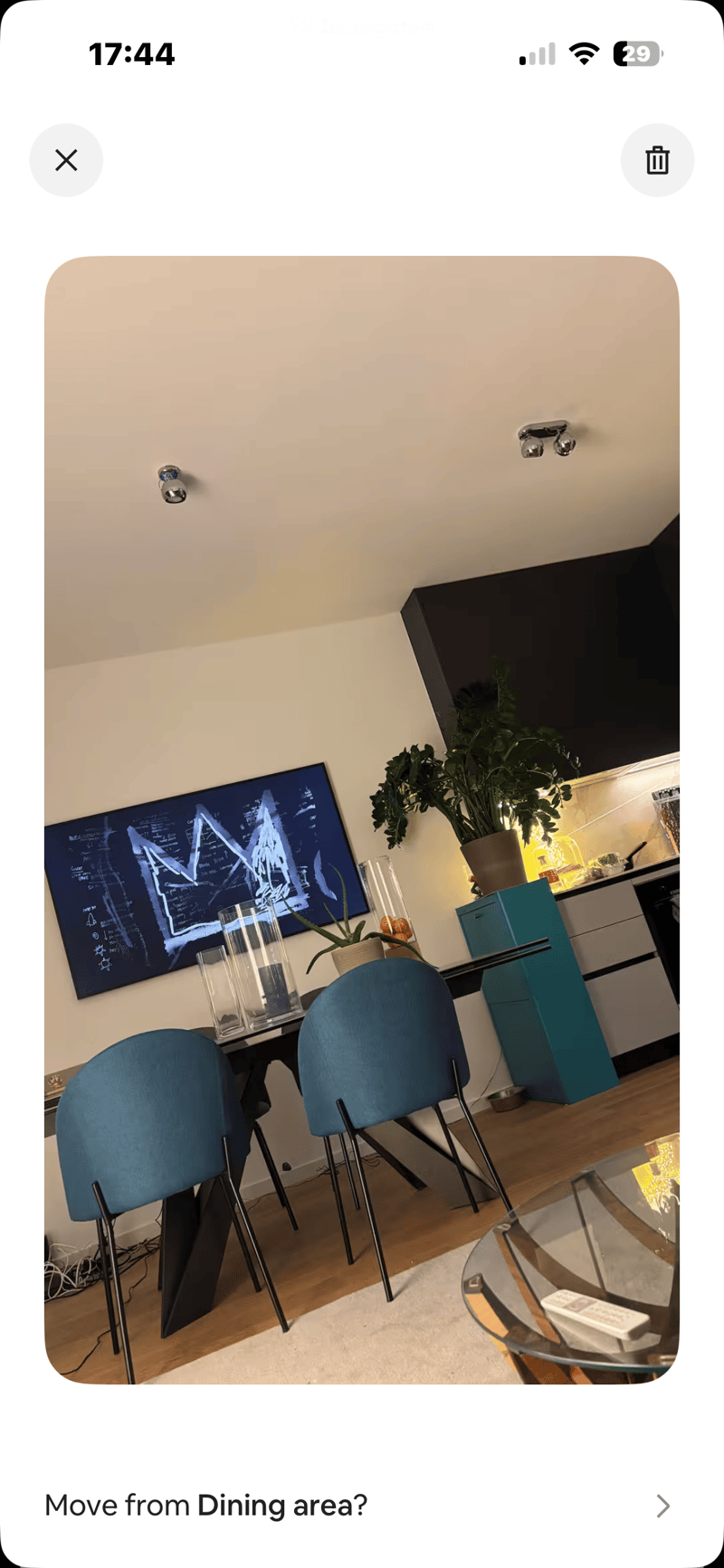 Dining area with a modern glass table, blue chairs, a potted plant, and a screen on the wall. The area is adjacent to a kitchen with cabinets and a countertop.