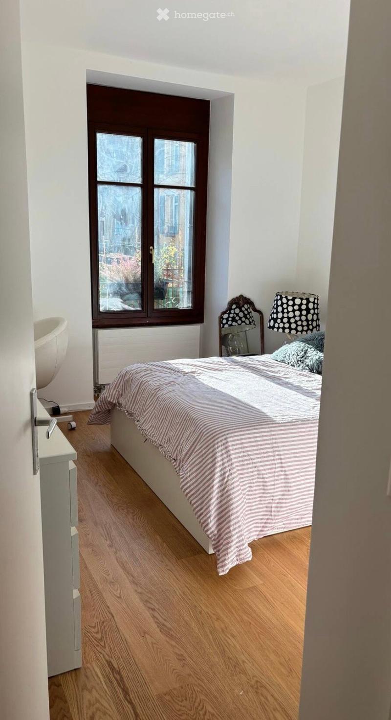 Cozy bedroom with wooden flooring, a double bed with striped bedding, two lamps on nightstands, and a white chair near the window.
