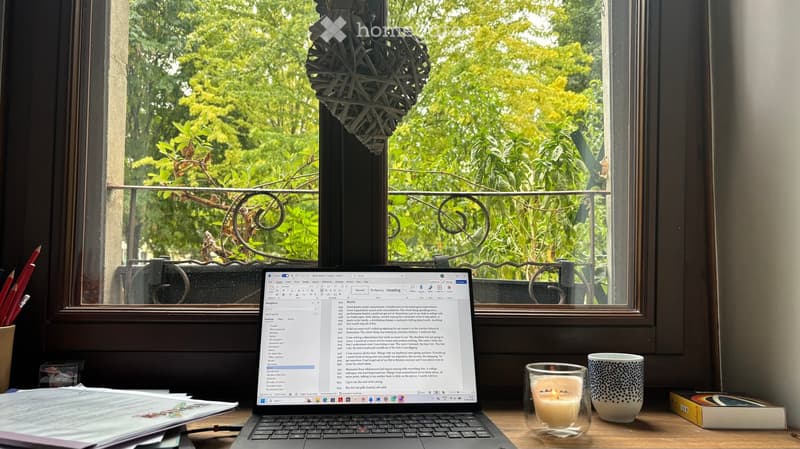 A cozy workspace with a laptop displaying a document, a lit candle, a cup, a book, and several pencils on a wooden desk, with a view of greenery through a large window.