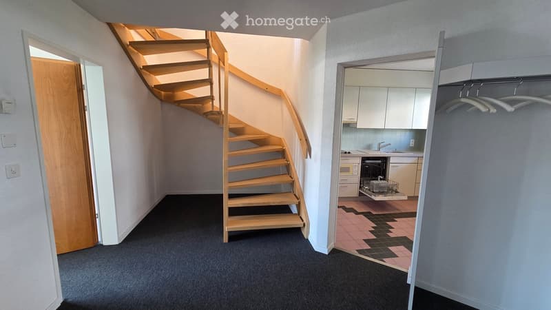 A modern hallway with a wooden staircase leading upwards, a doorway to a kitchen area with white cabinets, a dishwasher, and a tiled floor, and a coat rack with hangers on the right side.