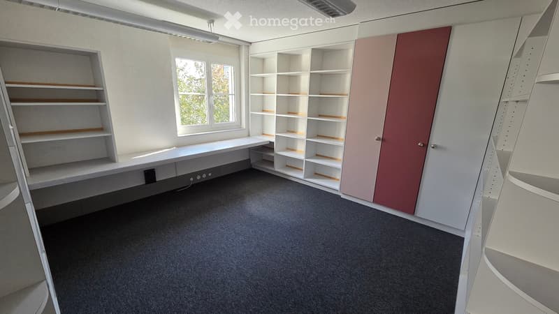 Empty room with built-in white shelving units, a desk, and a window with a view of greenery.