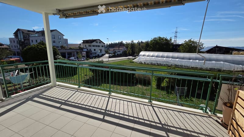Balcony with tiled floor, green metal railing, and a view of a residential area with houses, a greenhouse, and a clear blue sky.
