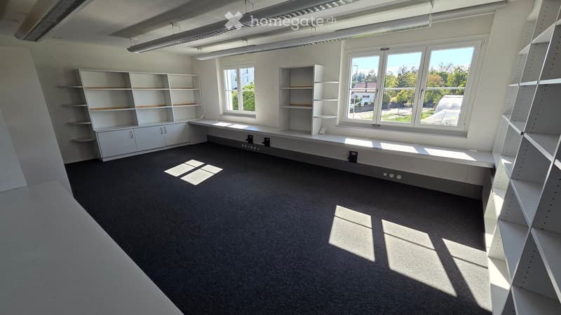 Empty room with white built-in shelving units, a long desk, and large windows allowing natural light to fill the space.