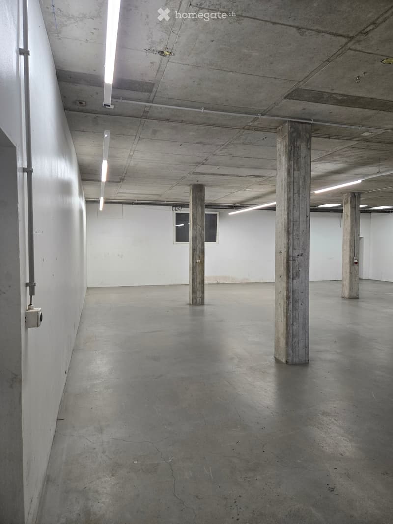 Empty industrial-style room with concrete floors, white walls, and exposed concrete pillars. Minimal lighting from fluorescent tubes and small windows.