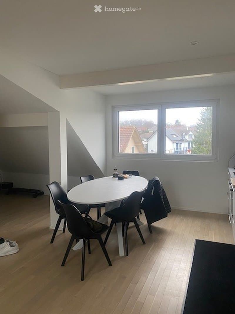 A modern dining area with a round white table and black chairs, wooden flooring, and large windows overlooking a residential neighborhood.