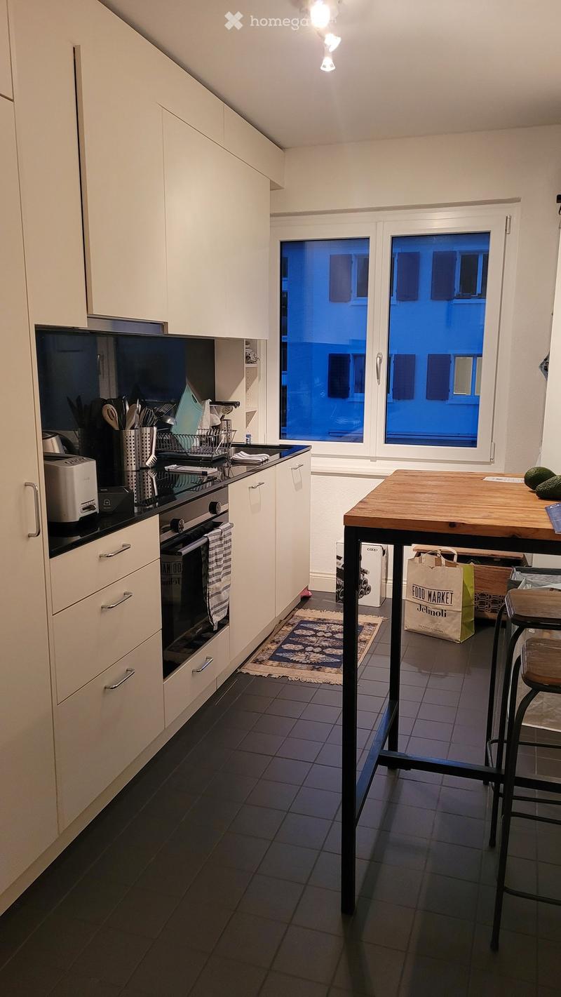 Modern kitchen with white cabinets, dark tiled floor, and a wooden island with a stool. Features an oven, toaster, and various kitchen utensils on the counter.