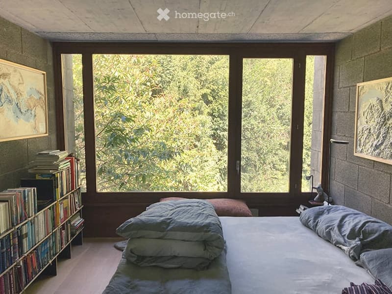 Bright bedroom with large windows offering a view of greenery, a neatly made bed with pillows and blankets, a bookshelf filled with books, and framed artwork on the walls.
