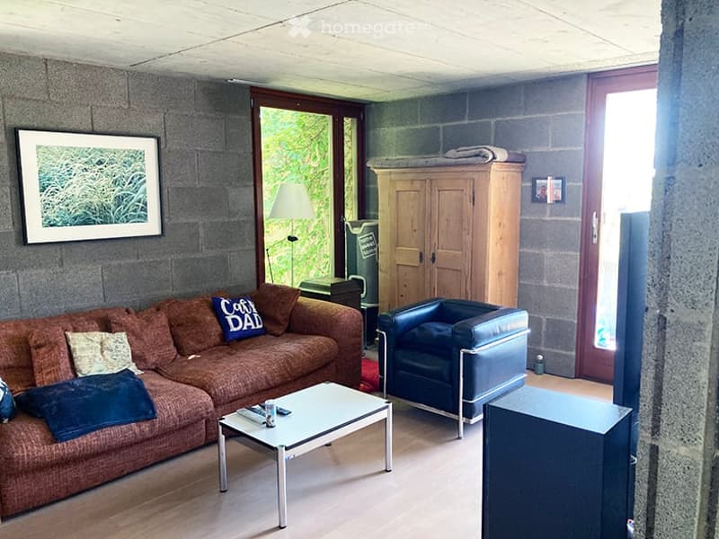 Living room with exposed stone walls, a brown sofa with throw pillows, a white coffee table, a blue armchair, a wooden cabinet, and a floor lamp. Large windows and a door lead to an outdoor area.