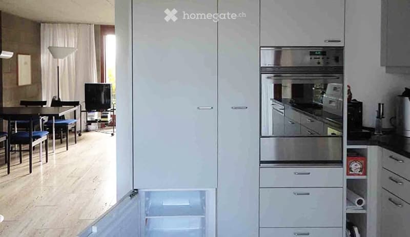 Modern kitchen with white cabinets, stainless steel appliances including a refrigerator, oven, and dishwasher. A dining area with a table and chairs is visible, along with a television and floor lamp in the background.