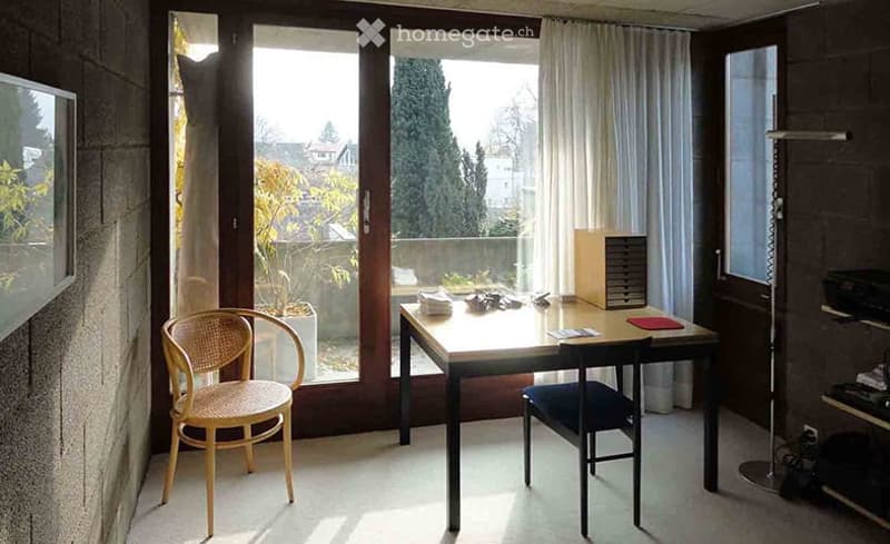 Bright room with large windows offering a view of greenery, furnished with a desk, two chairs, and a curtain.